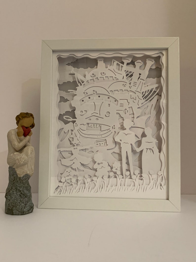 May include: A white paper cut-out art piece in a white frame depicting a flying castle with two figures standing in front of it. The castle is surrounded by clouds and flowers.