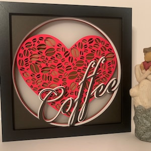 Könnte beinhalten: Ein gerahmtes 3D-Wandbild mit einer roten Herzform, die mit Kaffeekorn-Designs gefüllt ist, und dem Wort "Coffee" in einer stilisierten Schrift.