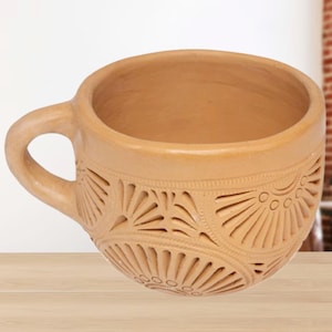 May include: A tan-colored ceramic teacup with a handle. The cup features intricate carved patterns around the exterior, including fan-like and circular designs. The cup sits on a light-colored wooden surface.