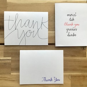 May include: Three white cards with different thank you messages. The first card has the words "thank you" written in black cursive. The second card has the words "merci", "tak", "thank you", "gracias", and "danke" written in black cursive. The third card has the words "Thank You" written in blue cursive.