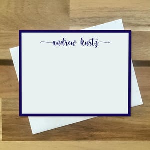 May include: A rectangular white stationery card with a thick dark blue border and the name "andrew kurtz" printed in dark blue cursive script at the top center, resting on top of a plain white envelope.