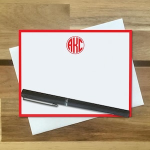 May include: A rectangular white note card with a bold red border and a red circular monogram featuring the letters AKC at the top center. A black ballpoint pen with a silver clip is positioned diagonally across the bottom portion of the card.