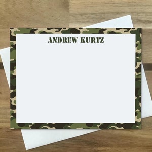 May include: A stationery set featuring a white card with a camouflage border and the name "ANDREW KURTZ" printed in green. The card is placed on top of a white envelope, set against a wooden background.