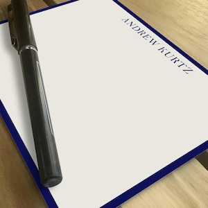 Personalized notepad with blue border on a wooden table. "Andrew Kurtz" printed in uppercase on the top right corner. Black pen placed diagonally on the blank surface. Ideal for professional or personal use, custom stationery, gift idea.