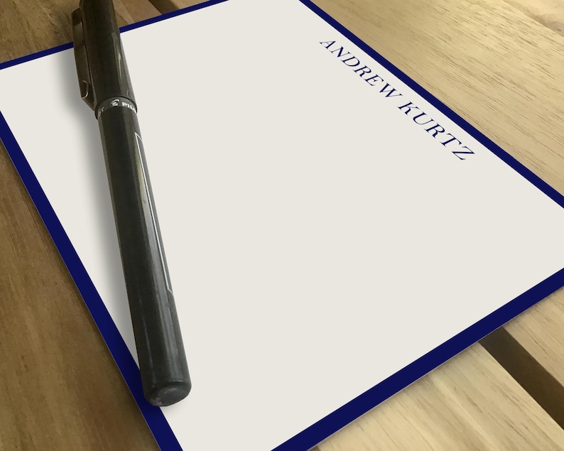 Personalized notepad with blue border on a wooden table. "Andrew Kurtz" printed in uppercase on the top right corner. Black pen placed diagonally on the blank surface. Ideal for professional or personal use, custom stationery, gift idea.