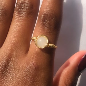 May include: A gold-colored wire-wrapped ring featuring a round, off-white stone. The ring is set on a finger, showcasing the delicate wirework and the smooth texture of the stone. The ring's design is simple yet elegant.