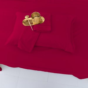 May include: A deep crimson red fitted sheet and two pillowcases. The sheet is folded and the pillowcases are stacked on top of the sheet. A gold metal tray with a gold spoon and a gold cup are on top of the pillowcases.