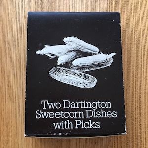 May include: A black and white box with a photo of two glass sweetcorn dishes with corn on the cob inside. The text on the box reads "Two Dartington Sweetcorn Dishes with Picks".
