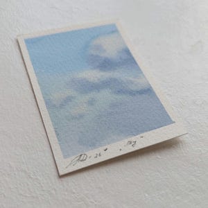 Pu&ograve; includere: Un piccolo acquerello di un cielo con nuvole. L'opera presenta sfumature di blu e bianco, con la parola "sky" scritta in corsivo. Firmato "AD 26".