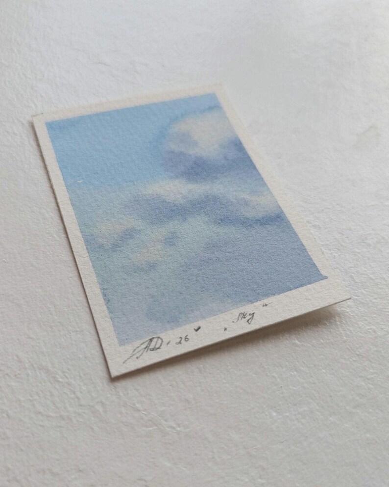 Pu&ograve; includere: Un piccolo acquerello di un cielo con nuvole. L'opera presenta sfumature di blu e bianco, con la parola "sky" scritta in corsivo. Firmato "AD 26".
