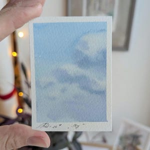 Pu&ograve; includere: Un piccolo dipinto ad acquerello di un cielo con nuvole. L'opera d'arte ha una forma rettangolare con uno sfondo azzurro chiaro e nuvole bianche e azzurre. La firma dell'artista e la parola "sky" sono visibili.
