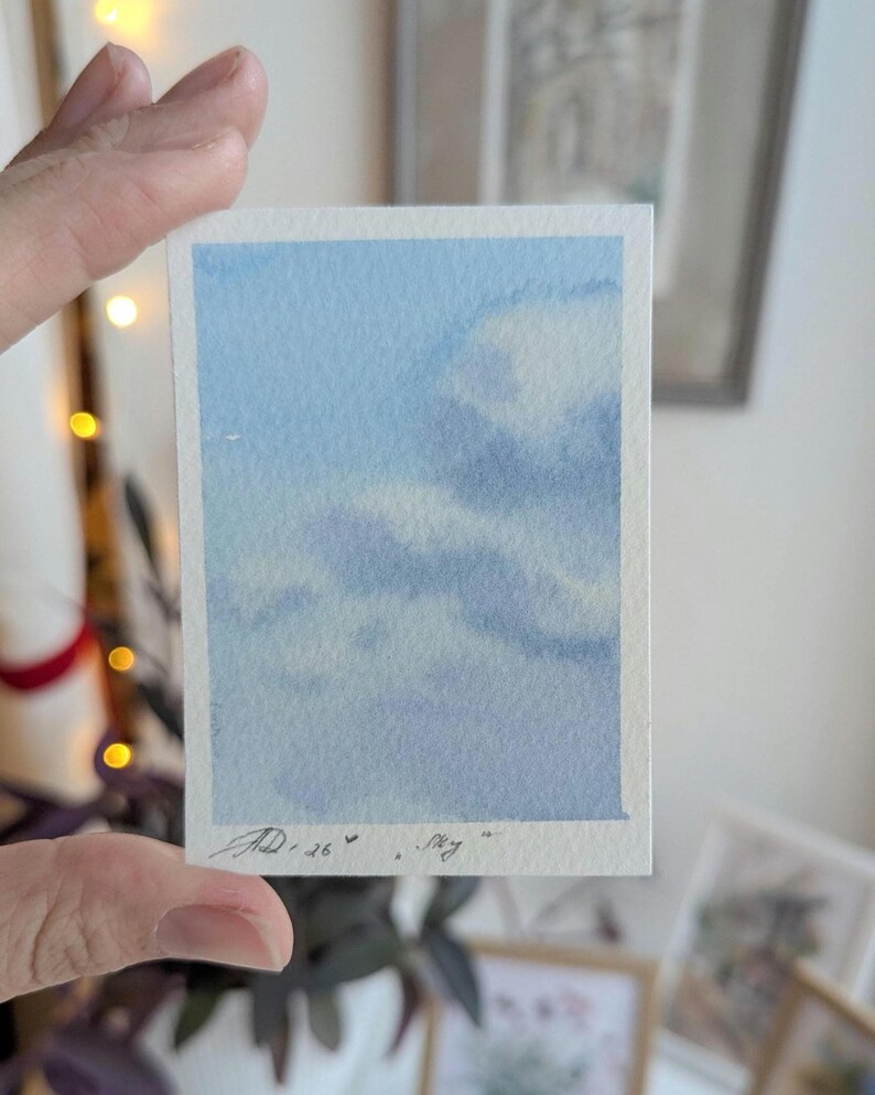 Pu&ograve; includere: Un piccolo dipinto ad acquerello di un cielo con nuvole. L'opera d'arte ha una forma rettangolare con uno sfondo azzurro chiaro e nuvole bianche e azzurre. La firma dell'artista e la parola "sky" sono visibili.