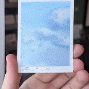 Pu&ograve; includere: Un piccolo acquerello di un cielo blu con nuvole bianche. L'opera d'arte &egrave; su un pezzo di carta rettangolare con un bordo bianco. La firma e la data dell'artista sono nell'angolo in basso a sinistra.