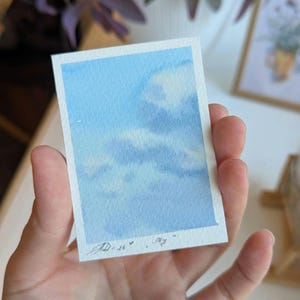 Pu&ograve; includere: Un piccolo acquerello di un cielo blu con nuvole bianche. L'opera d'arte &egrave; tenuta in mano, mostrando le delicate pennellate e le morbide transizioni di colore. Firmato "AD .26" con la parola "sky" sotto.