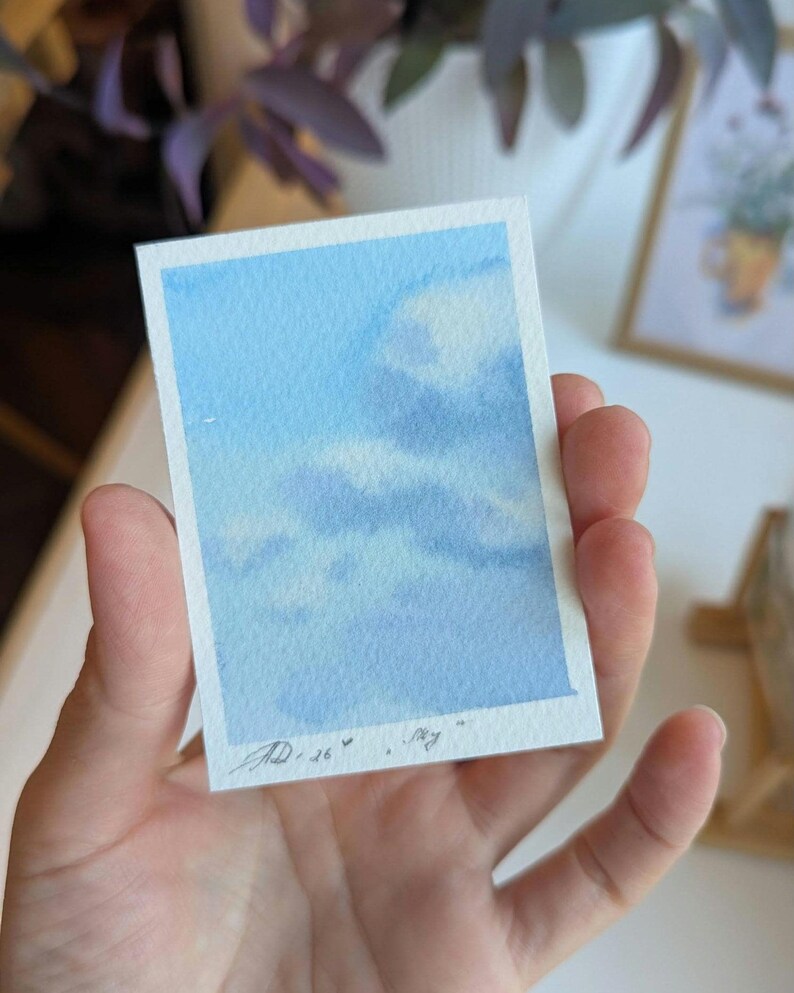 Pu&ograve; includere: Un piccolo acquerello di un cielo blu con nuvole bianche. L'opera d'arte &egrave; tenuta in mano, mostrando le delicate pennellate e le morbide transizioni di colore. Firmato "AD .26" con la parola "sky" sotto.