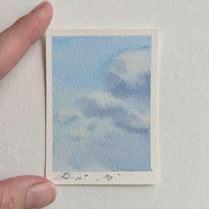 Pu&ograve; includere: Un piccolo acquerello di un cielo blu con nuvole bianche. L'opera d'arte &egrave; su un pezzo di carta rettangolare con un bordo bianco. La firma dell'artista e la parola "Sky" sono visibili nell'angolo in basso a destra.