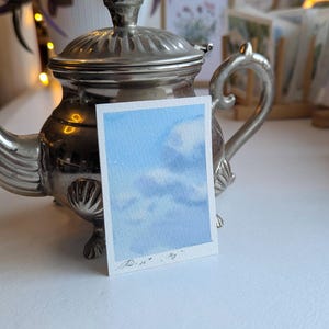 Pu&ograve; includere: Un piccolo acquerello di un cielo blu con nuvole bianche. L'opera d'arte &egrave; su un pezzo di carta rettangolare, tenuto davanti a una teiera d'argento. Il dipinto &egrave; firmato in basso a sinistra.