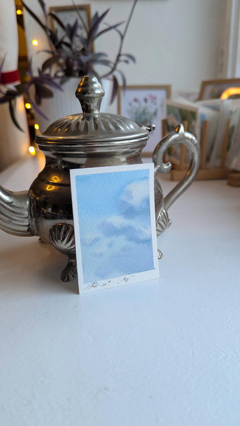 Pu&ograve; includere: Un piccolo acquerello di un cielo blu con nuvole bianche. L'opera d'arte &egrave; su un pezzo di carta rettangolare, tenuto davanti a una teiera d'argento. Il dipinto &egrave; firmato in basso a sinistra.