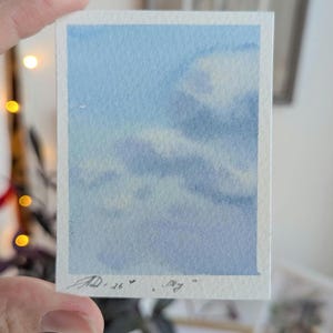 Pu&ograve; includere: Un piccolo acquerello di un cielo blu con soffici nuvole bianche. L'opera d'arte &egrave; firmata "AD.26" e include la parola "Sky". Il dipinto &egrave; su un foglio di carta rettangolare con un bordo bianco.