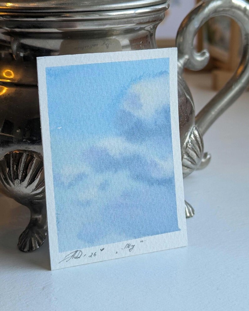 Pu&ograve; includere: Un piccolo acquerello di un cielo blu con soffici nuvole bianche. L'opera d'arte &egrave; firmata e datata "JD '26" con la parola "Sky" scritta sotto. Il dipinto &egrave; su una carta rettangolare bianca.