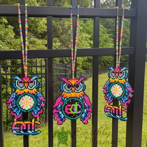 Puede incluir: Tres collares de cuentas con forma de búho de colores que cuelgan de una valla de metal negro. Cada búho tiene un diseño diferente, incluyendo una flor, un círculo con las letras "ETC" y un patrón geométrico. Los collares están hechos de pequeñas cuentas de colores.