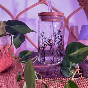 May include: A clear glass jar with a black floral design and a bamboo lid. The jar has a glass straw sticking out of the lid. The jar is sitting on a book with a plant in the background.