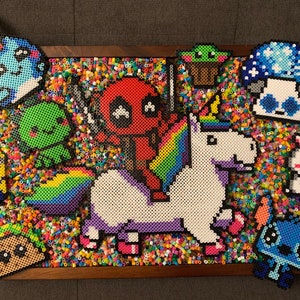 May include: A colorful beaded art piece featuring a unicorn with a rainbow mane and tail, a red and black Deadpool character riding on its back, and other characters like a green alien, a blue mushroom, and a yellow minion. The background is a mix of colorful beads.