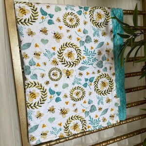 May include: A white baby blanket featuring a pattern of yellow and black bees, green leaves, and yellow flowers. The blanket has a teal, plush backing. The blanket is draped over a gold-coloured rack.