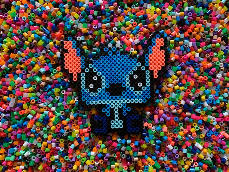 May include: A blue, orange, and black pixel art character resembling Stitch from Lilo & Stitch, surrounded by colorful plastic beads.