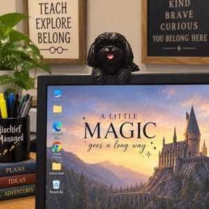 May include: A black Shih Tzu dog figurine with a textured coat and a pink tongue, perched on the top edge of a computer monitor. The monitor displays a desktop background featuring a castle and the text A LITTLE MAGIC goes a long way.