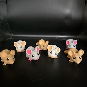 Puede incluir: Una colección de siete pequeñas figuritas de ratón grises y marrones. Los ratones tienen orejas rosas y ojos negros. Están dispuestos en semicírculo sobre una superficie negra.