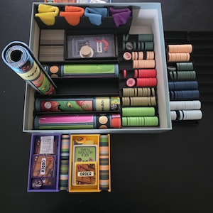 May include: A board game organizer with various components. The box contains cards, tokens, and rolled-up playmats. The components are organized in trays and include colorful game pieces and cards with the word "ORDER" on them.