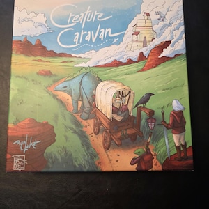 May include: A board game box titled "Creature Caravan" featuring a fantasy landscape. The artwork depicts a dinosaur pulling a covered wagon along a path, with a castle in the background. A person with a sword stands nearby.
