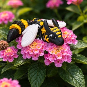 May include: A whimsical, black, yellow, and white bee-dragon hybrid figurine rests on a cluster of pink flowers with green leaves. The bee-dragon has white wings and a detailed, stylized design. The background is blurred, emphasizing the figurine.