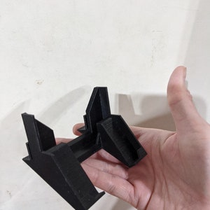 Könnte beinhalten: Ein schwarzer 3D-gedruckter Ständer mit geometrischem Design. Der Ständer hat eine flache Basis und drei abgewinkelte Stützen.
