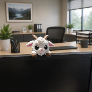 May include: A small, white, 3D-printed goat figurine with brown horns and hooves, large black eyes, and pink inner ears. The goat has a tuft of white hair on its head and is positioned to appear as if it is peering over the top edge of a black monitor.