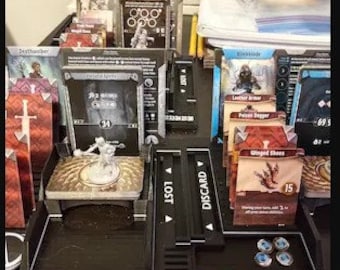 Pode incluir: Uma configuração de jogo de tabuleiro com uma variedade de cartas, incluindo cartas de personagem, cartas de itens e uma pilha de descarte. As cartas estão dispostas em uma superfície preta com um botão vermelho e um conjunto de botões numerados.