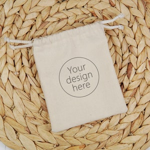 以下が含まれることがあります： 「Your design here」の文字が円で囲まれたベージュ色の巾着袋。天然繊維で織られた円形のプレースマットの上に置かれています。背景は白い表面です。