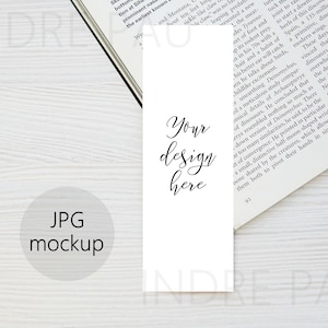 May include: A white bookmark mockup with the text "Your design here" written in black cursive script. The bookmark is laying on a white wooden surface with a book partially visible in the background.