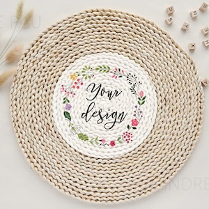 Könnte beinhalten: Ein runder geflochtener Platzteller mit einem weißen Kreis in der Mitte. Der Kreis hat ein florales Kranzdesign mit dem Text "Your design" in der Mitte.