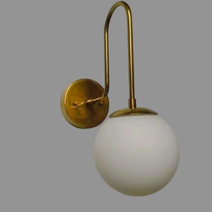 May include: A wall-mounted light fitting with a brass finish and a white globe shade. The fitting has a curved arm and a circular base for mounting. The globe is a smooth, opaque white.