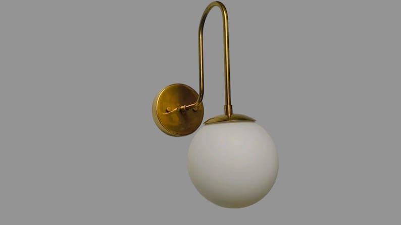 May include: A wall-mounted light fitting with a brass finish and a white globe shade. The fitting has a curved arm and a circular base for mounting. The globe is a smooth, opaque white.