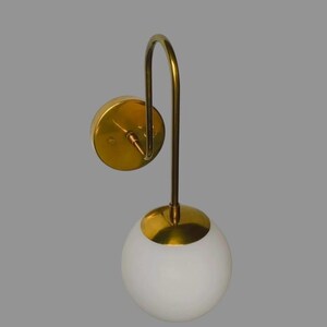 May include: A contemporary wall light with a gold finish and a white globe shade. The sconce features a circular wall mount, a curved arm, and a spherical light fixture. The design is minimalist and elegant, suitable for various interior styles.