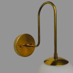 May include: A brass wall light with a curved arm and a circular backplate. It features a white, globe-shaped glass shade. The light fixture is designed for indoor use.