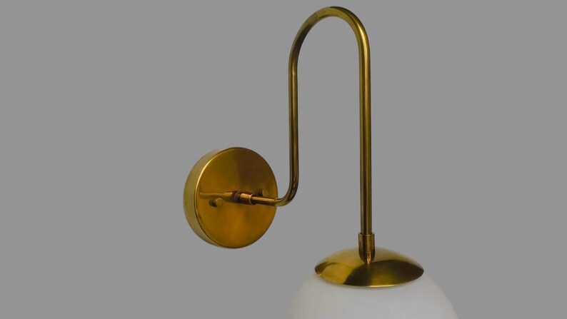 May include: A brass wall light with a curved arm and a circular backplate. It features a white, globe-shaped glass shade. The light fixture is designed for indoor use.
