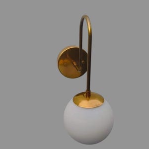 May include: A contemporary wall light with a white globe shade and a curved, brass-coloured arm. The light has a circular base and a matching brass-coloured cap. The fixture is designed for wall mounting and provides ambient lighting.