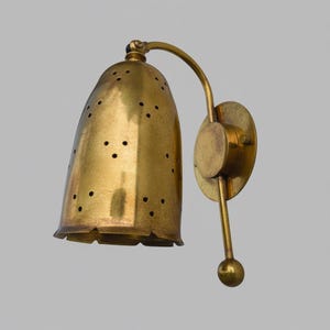 May include: A brass wall sconce featuring a bell-shaped shade with a perforated pattern of small circular holes. The fixture includes a curved arm, a circular wall mounting plate, and a vertical rod ending in a small spherical finial at the base.