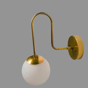 May include: A contemporary wall light featuring a gold-toned metal frame and a spherical, frosted glass shade. The fixture has a curved arm and a circular wall plate. The design is minimalist and suitable for various interior styles.