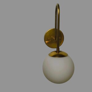 May include: A wall-mounted light fitting with a gold-coloured metal frame and a white, spherical glass shade. The fitting has a circular base and a curved arm. The shade is approximately 15 cm in diameter.