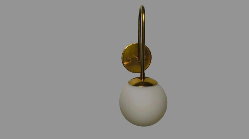 May include: A wall-mounted light fitting with a gold-coloured metal frame and a white, spherical glass shade. The fitting has a circular base and a curved arm. The shade is approximately 15 cm in diameter.